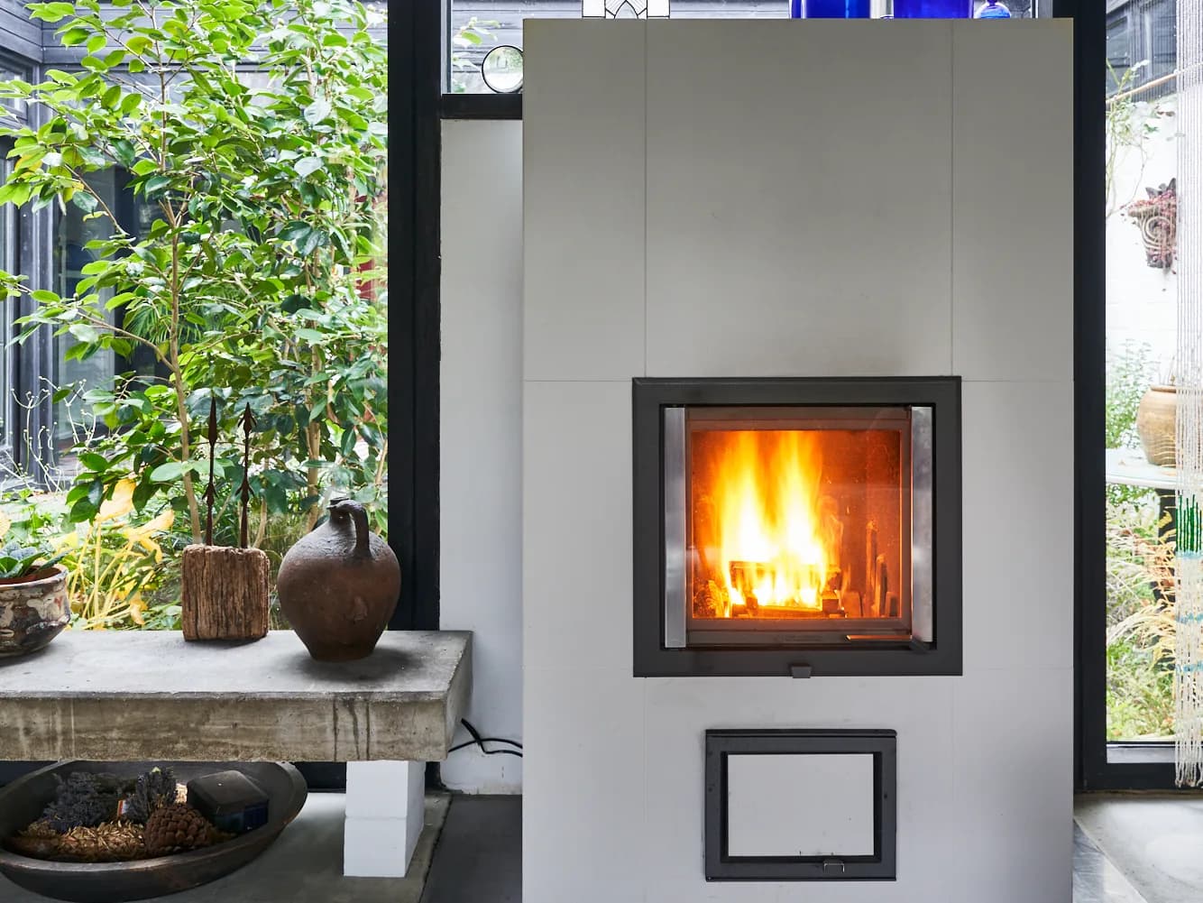 Specksteinofen mit heller Keramikverkleidung und brennendem Feuer in einem verglasten Raum, daneben eine Steinbank und Grünpflanzen